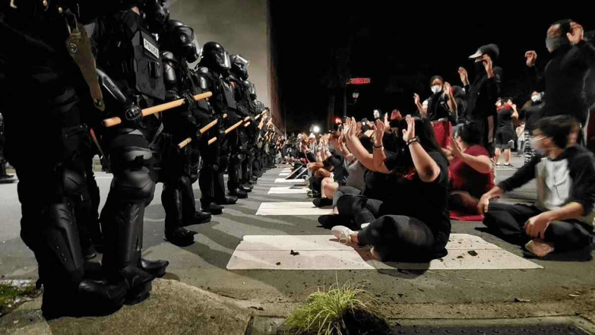 A line of police in riot gear facing seated protesters with raised hands at a nighttime demonstration in Raleigh.